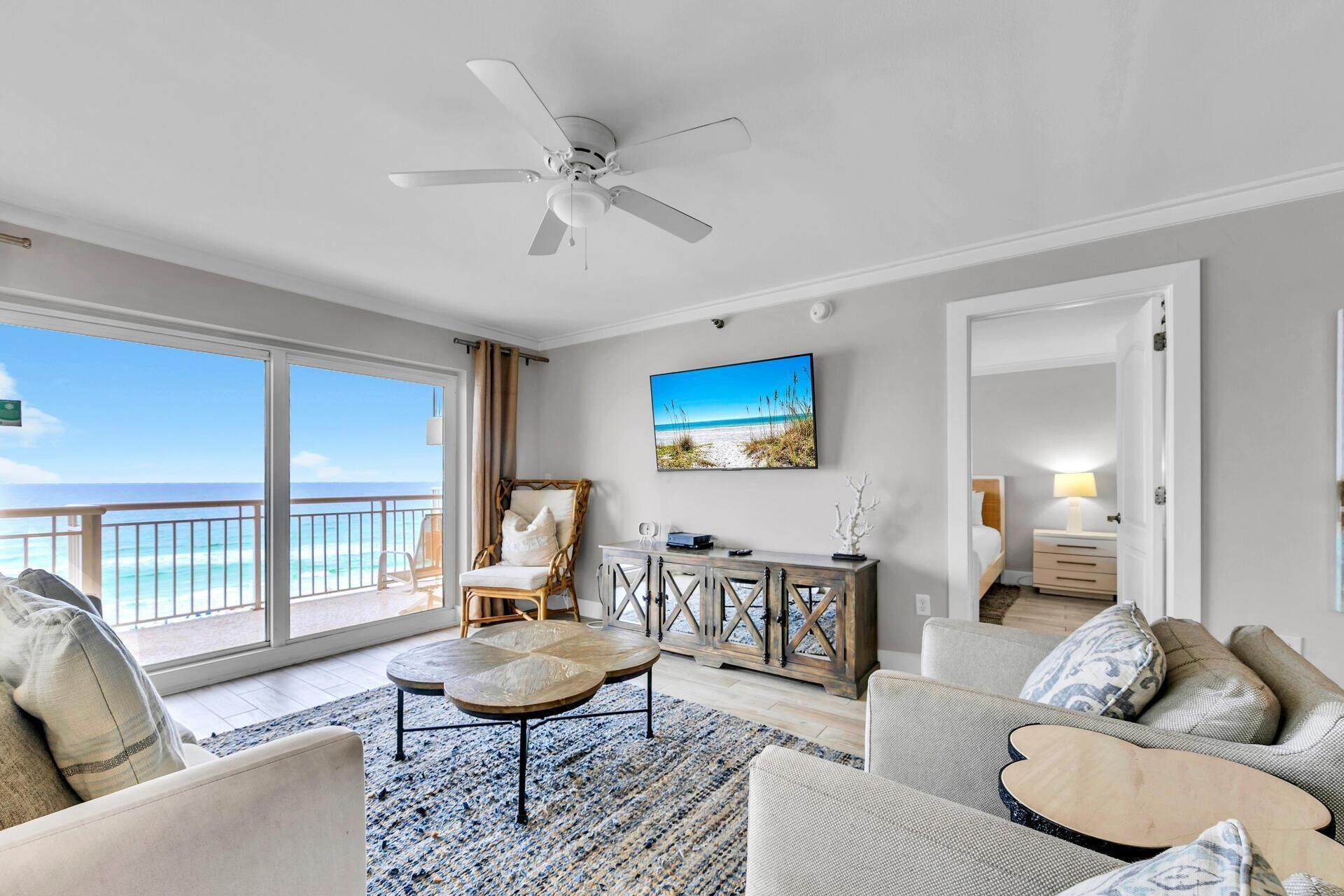 A spacious living room with a large balcony, a ceiling fan, a mounted TV, and a wooden cabinet.