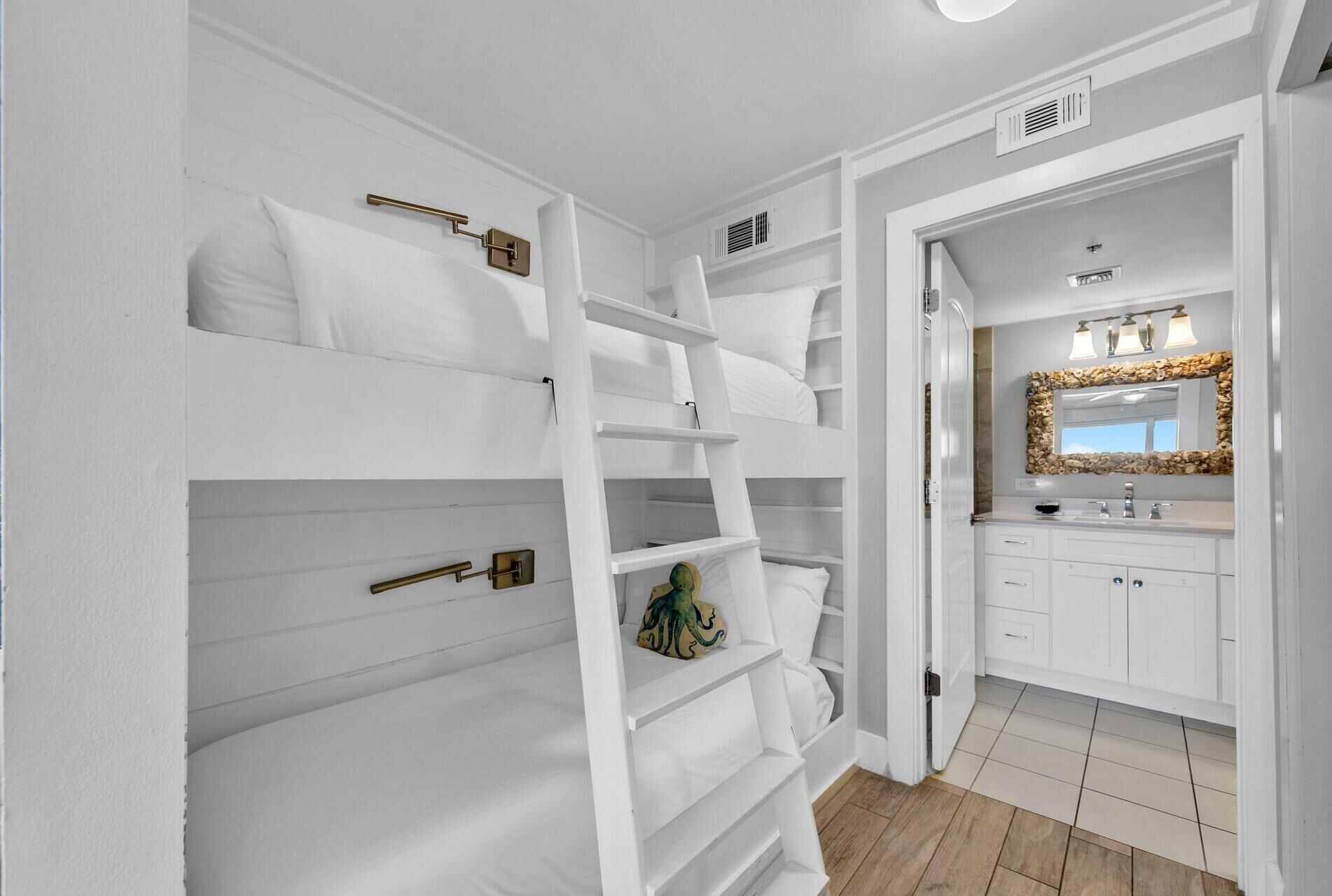 Ambiente interno bianco con una cuccetta, un bagno con lavandino e specchio, e un pavimento in legno.