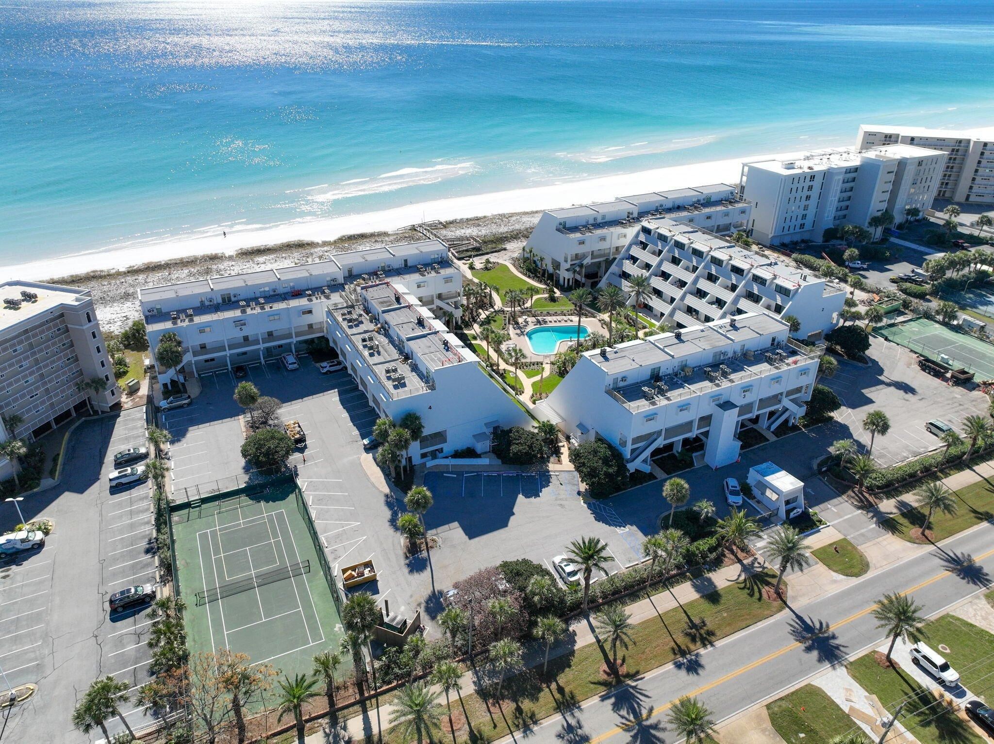 Una vista aerea di un complesso alberghiero costiero con diversi edifici, un campo da tennis e una piscina.