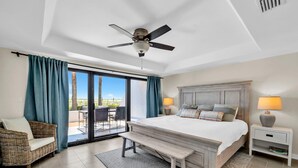 Un dormitorio con una cama grande, ventilador de techo, una banca, mesita de noche y vistas al exterior.