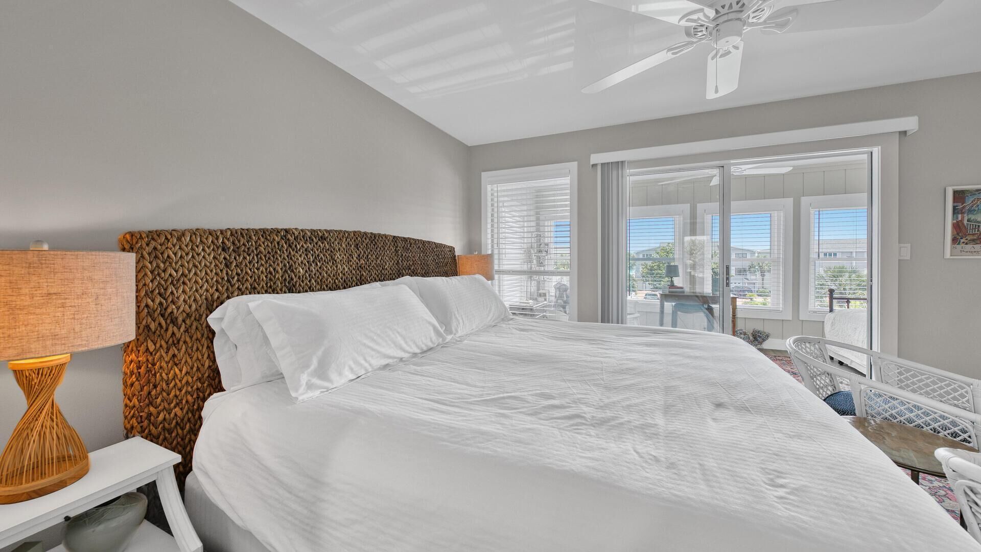 Quarto com cama, luminária, ventilador de teto e vista para o exterior através de janelas amplas.