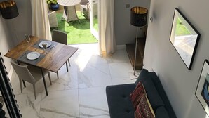 A dining area with a table and chairs, a sofa, and a view of a garden through a glass door. - Wimbledon - Apartment - Sleeps 2 -private Entrance (London)