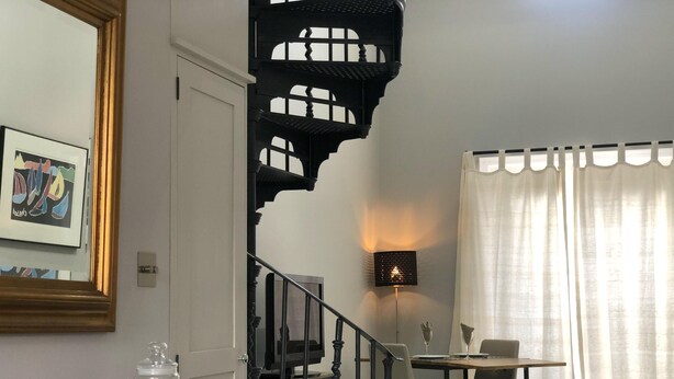 A dining area with a table and chairs, a staircase with a black railing, a wall-mounted lamp, and a window with curtains. - Wimbledon - Apartment - Sleeps 2 -private Entrance (London)