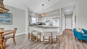 A modern kitchen with white cabinets, a central island with stools, a dining table with chairs, and a blue sofa. - Beach Bum Hideaway (Santa Rosa Beach)