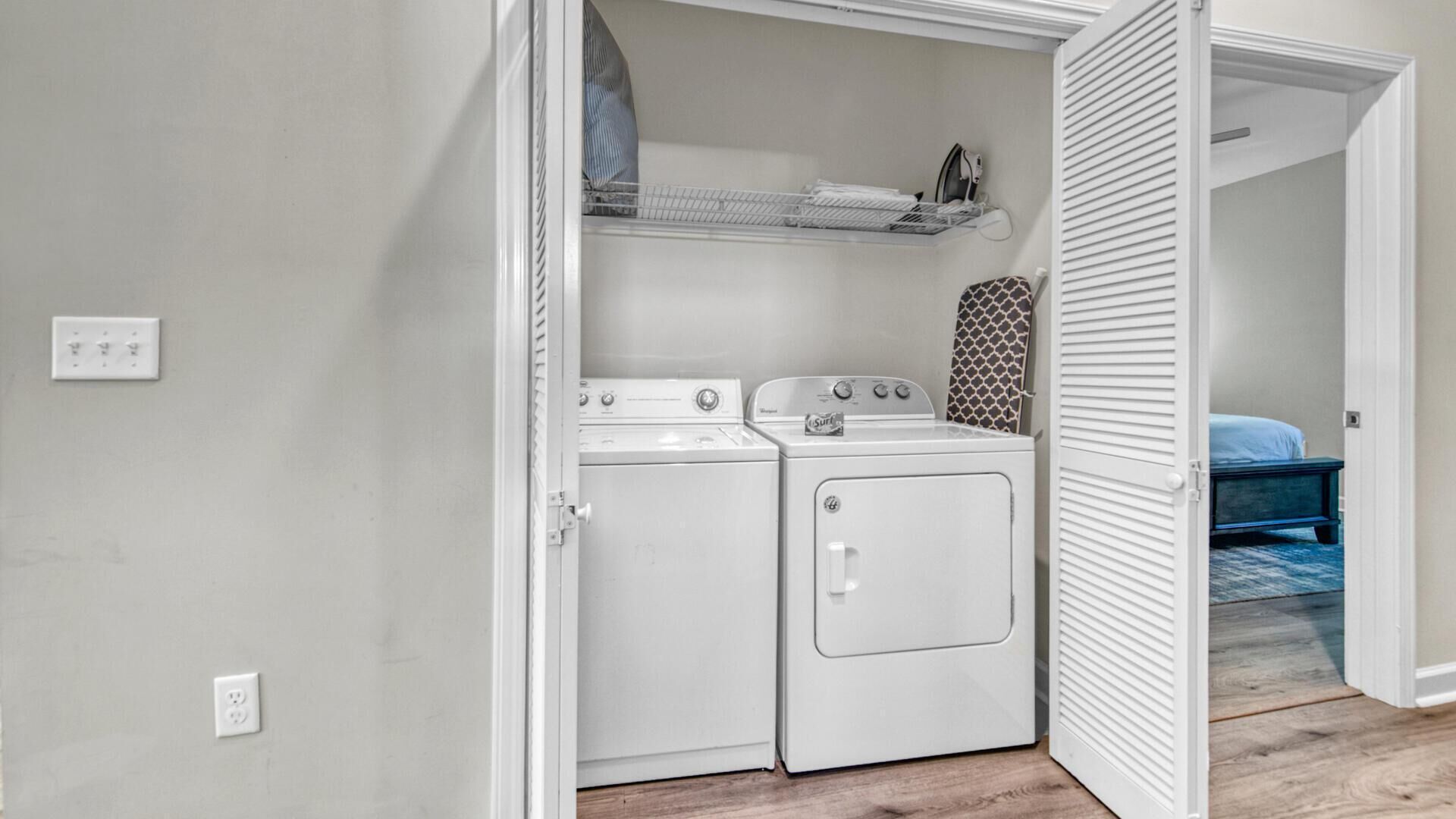 A laundry room with a washer and dryer, a shelf above the dryer, and a bed in the adjacent room.