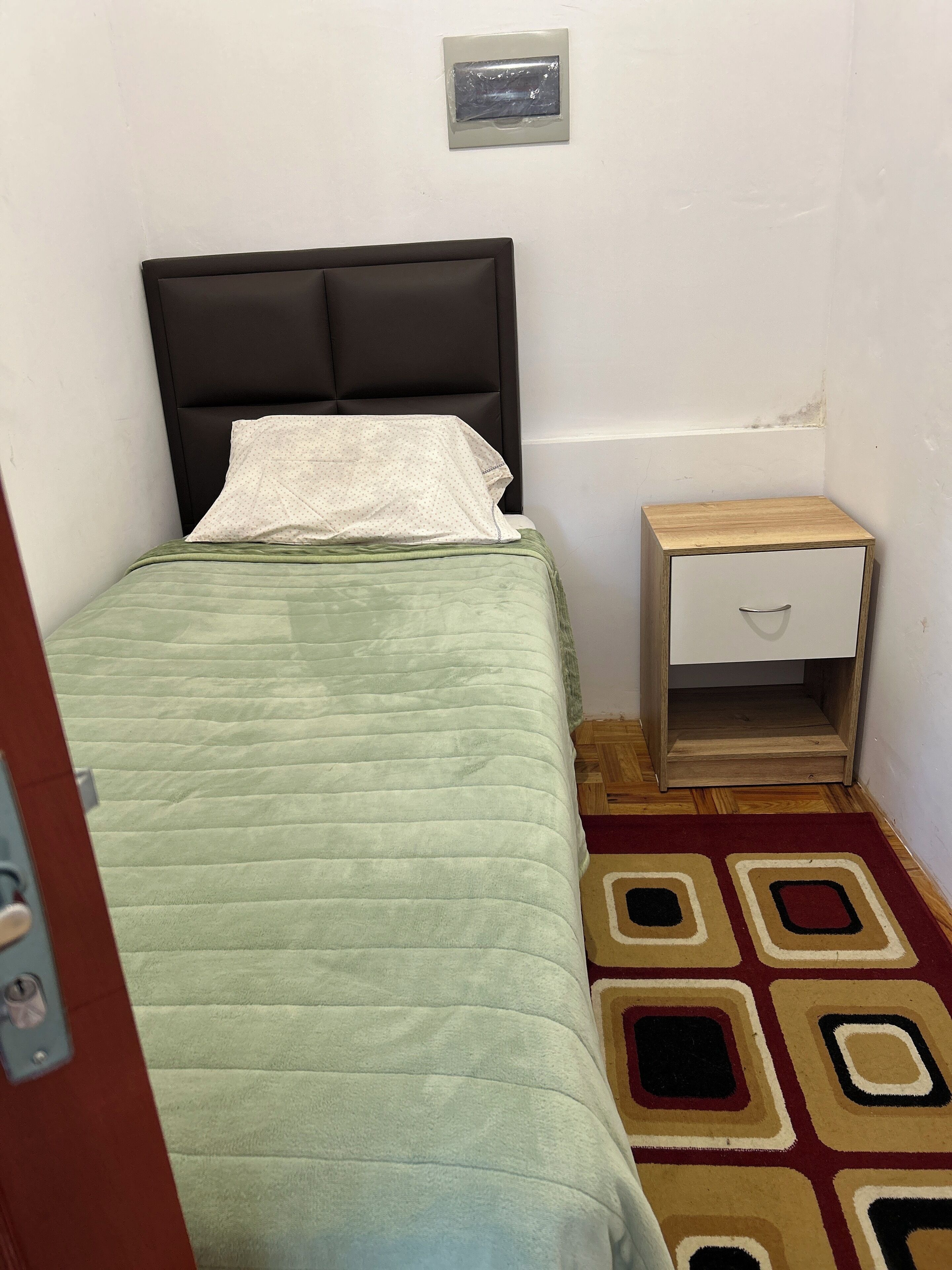 A small, single-bed room with a green quilt, a wooden nightstand with a drawer, and a patterned rug.