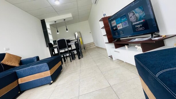A modern living room with a flat-screen TV displaying streaming services, a sofa, a dining area, and a kitchen in the background. - Apartamento Nuevo, Moderno, Acogedor y muy Accesible a Todo en San Salvador (Mejicanos)