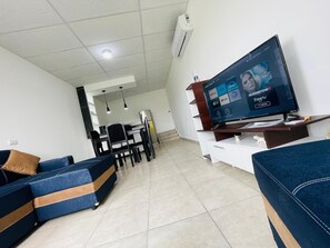 Un salon moderne doté d’un téléviseur à écran plat diffusant des services de streaming, d’un canapé, d’un coin repas et d’une cuisine en arrière-plan.