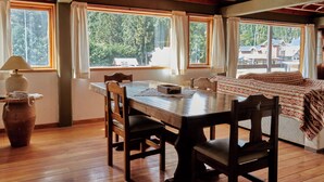 Une table de salle à manger en bois, avec des chaises, un vase et une lampe. Il y a des fenêtres avec des rideaux, un lit recouvert d’une couverture à motifs, et on peut voir des maisons et des arbres à l’extérieur.