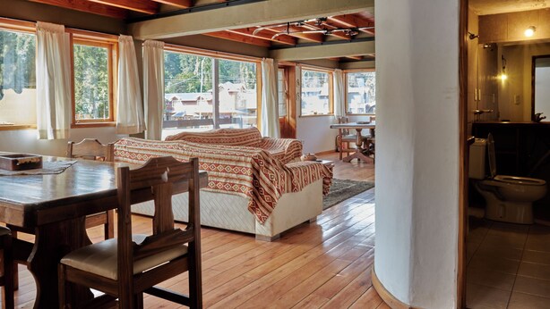 Une pièce chaleureuse avec un plancher en bois, un lit recouvert d’une literie à motifs, une table à manger avec des chaises et de grandes fenêtres donnant sur l’extérieur.