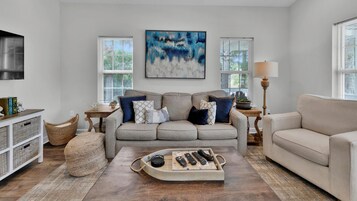 A living room with a grey sofa, a wooden coffee table with remote controls, a television on the left, and a painting above the sofa.