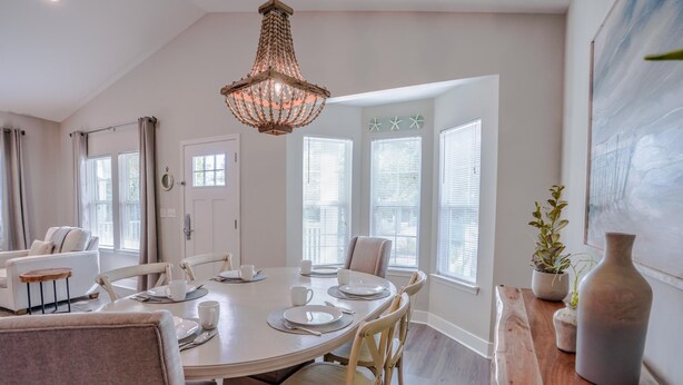 Uma sala de jantar com mesa redonda, cadeiras e um candelabro. Há janelas com persianas e uma porta.