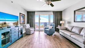 Uma sala de estar ampla com piso de madeira, um sofá, duas poltronas, uma TV e uma varanda com vista para a praia.