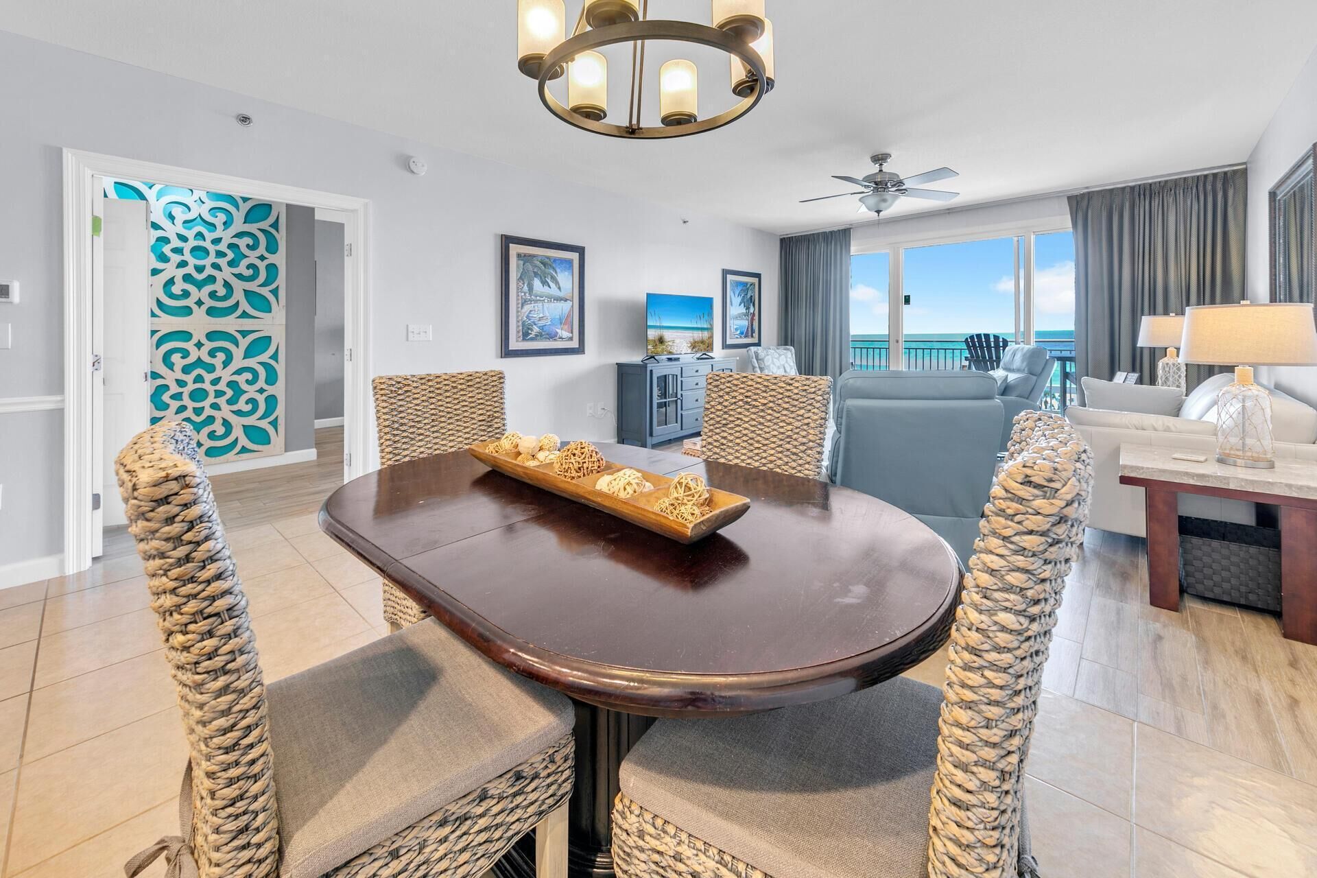 Een moderne hotelkamer met een zithoek, bestaande uit een houten tafel en geweven stoelen, een balkon met uitzicht op zee en een plafondventilator.