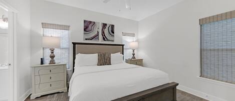 A bedroom with a bed, bedside tables, a ceiling fan, artwork on the wall, and a window with blinds.