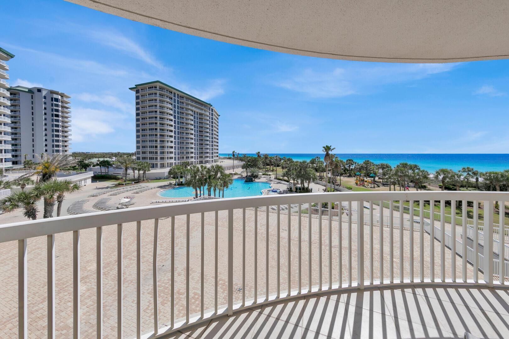 A balcony with a view of a swimming pool, palm trees, and a beach.