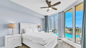 Habitación de hotel moderna con una cama grande, una mesita de noche, un ventilador de techo y vistas a una piscina y edificios altos.