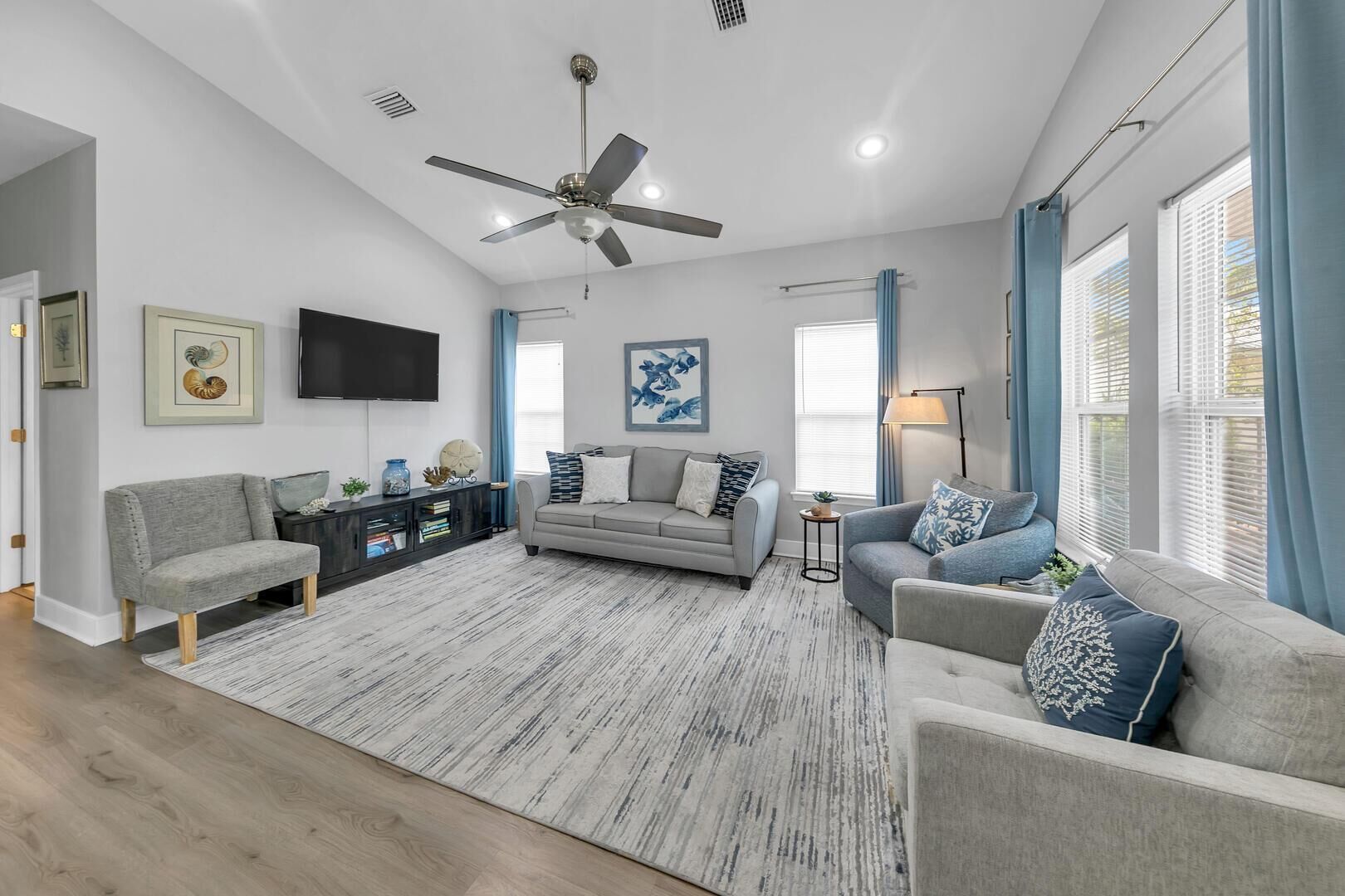 A spacious living room with a grey sofa, a grey armchair, and a coffee table. There is a ceiling fan, a flat-screen TV mounted on the wall, and a window with blue curtains.