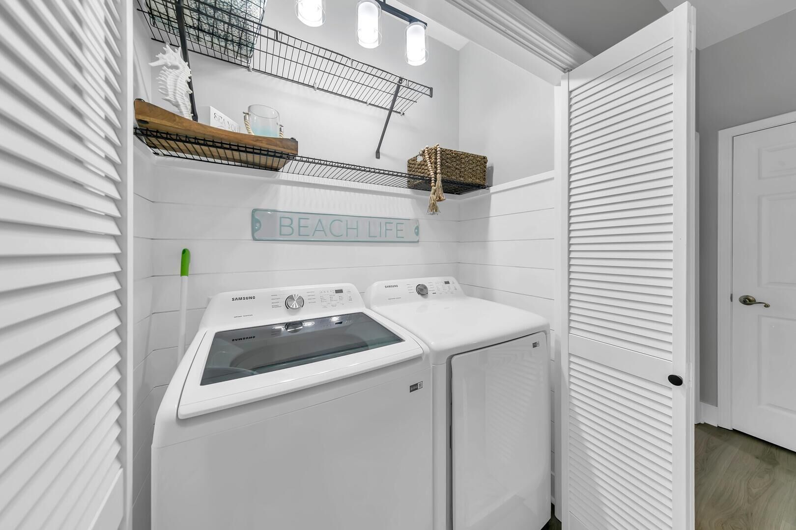 A laundry room with two white Samsung washing machines, a shelf with a towel and a basket, and a sign that reads "BEACH LIFE".