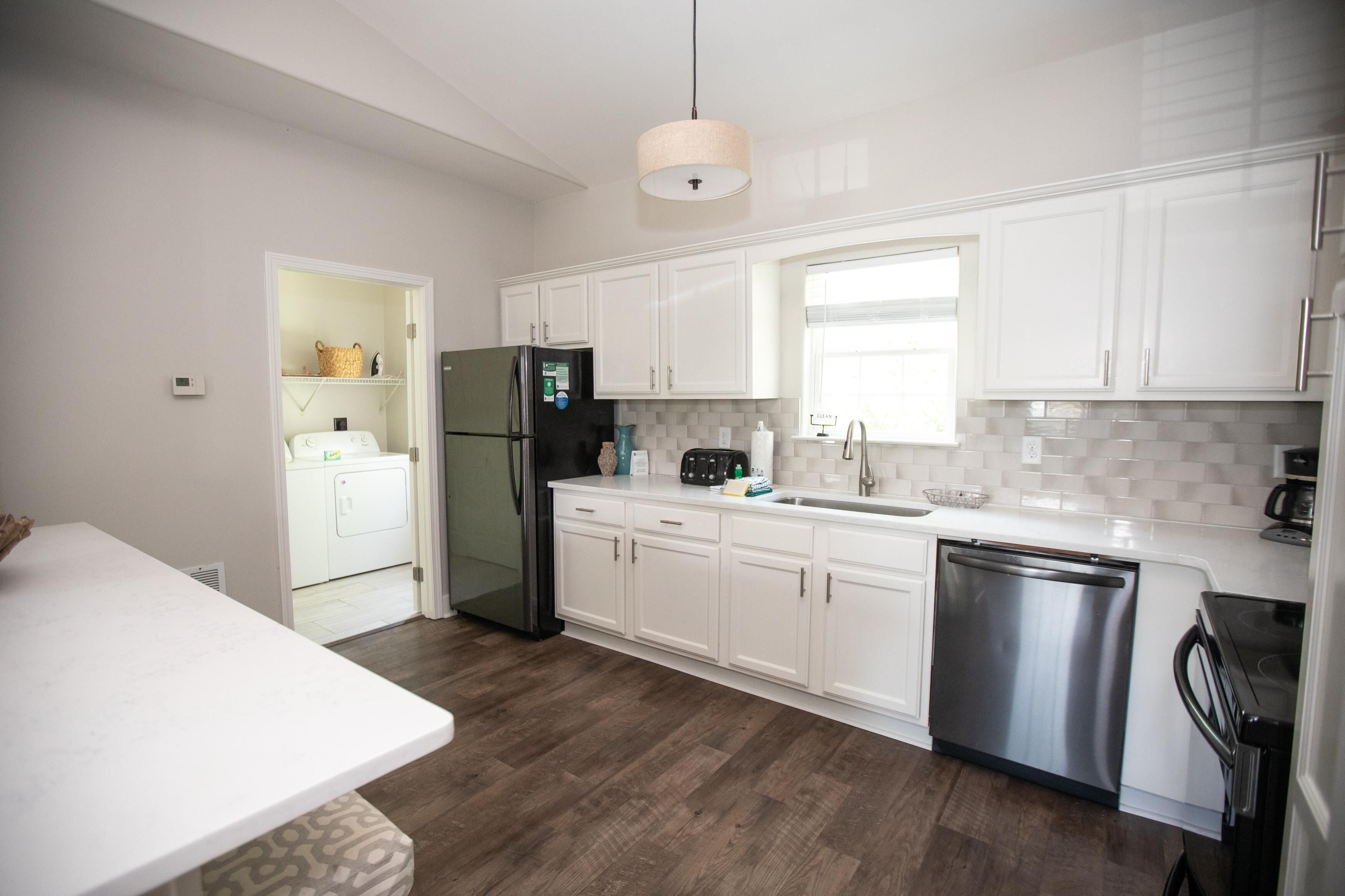 A modern kitchen with white cabinets, a stainless steel refrigerator, a dishwasher, and a window above the sink.