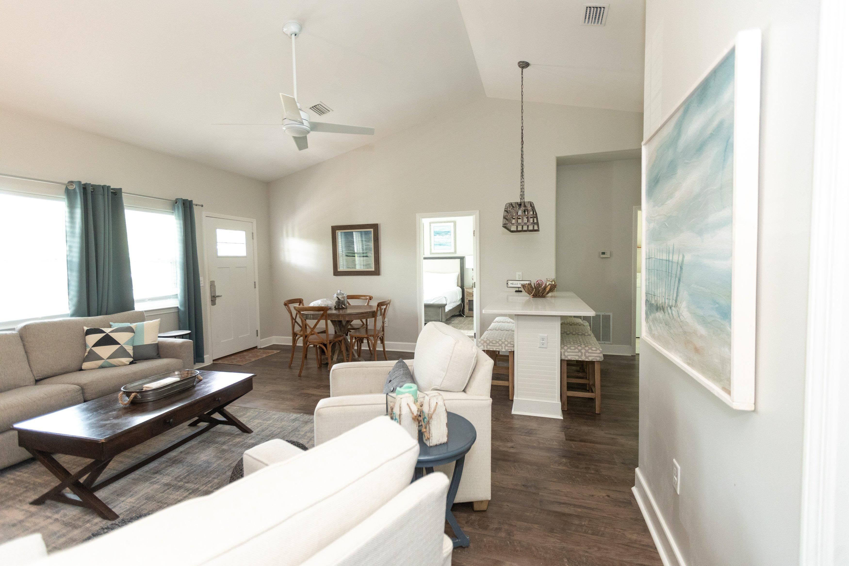 A living room with a sofa, coffee table, and chairs, a dining area with a table and chairs, and a kitchen area with a counter and hanging light fixture.