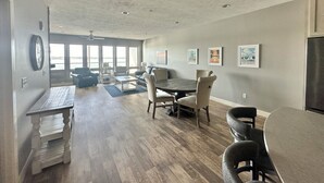 A spacious living room with a large dining table, comfortable seating, and a view of the outdoors through large windows. - Bayside Vacation Condo Rental 4 bedrooms!! (Arnolds Park)