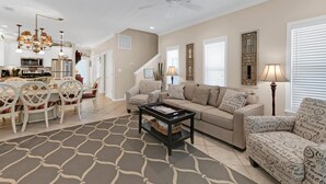 Ein geräumiges Wohnzimmer mit Sofa, Sesseln und Couchtisch. Es gibt einen Essbereich mit Stühlen und einer Chandelier. Im Hintergrund sind Treppe und Küche zu sehen.