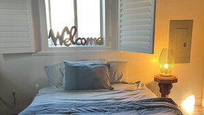 A bedroom with a bed, pillows, a nightstand with a lamp, a window with blinds, and a 'Welcome' sign. - Hidden gem. Tiny home in mural district.  (Blythe)