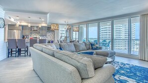 A modern living room with a large sectional sofa, dining area, and large windows offering a city view. - St. Maarten 701 at Silver Shells (Destin)