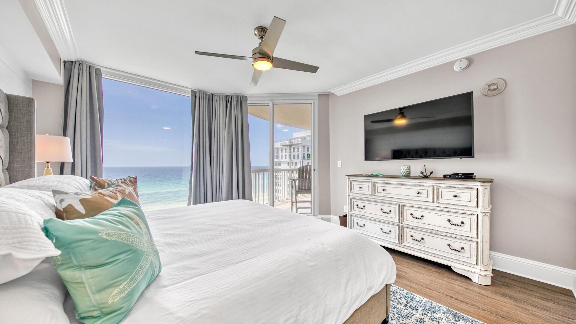 A bedroom with a large bed, a ceiling fan, a TV, and a view of the ocean.