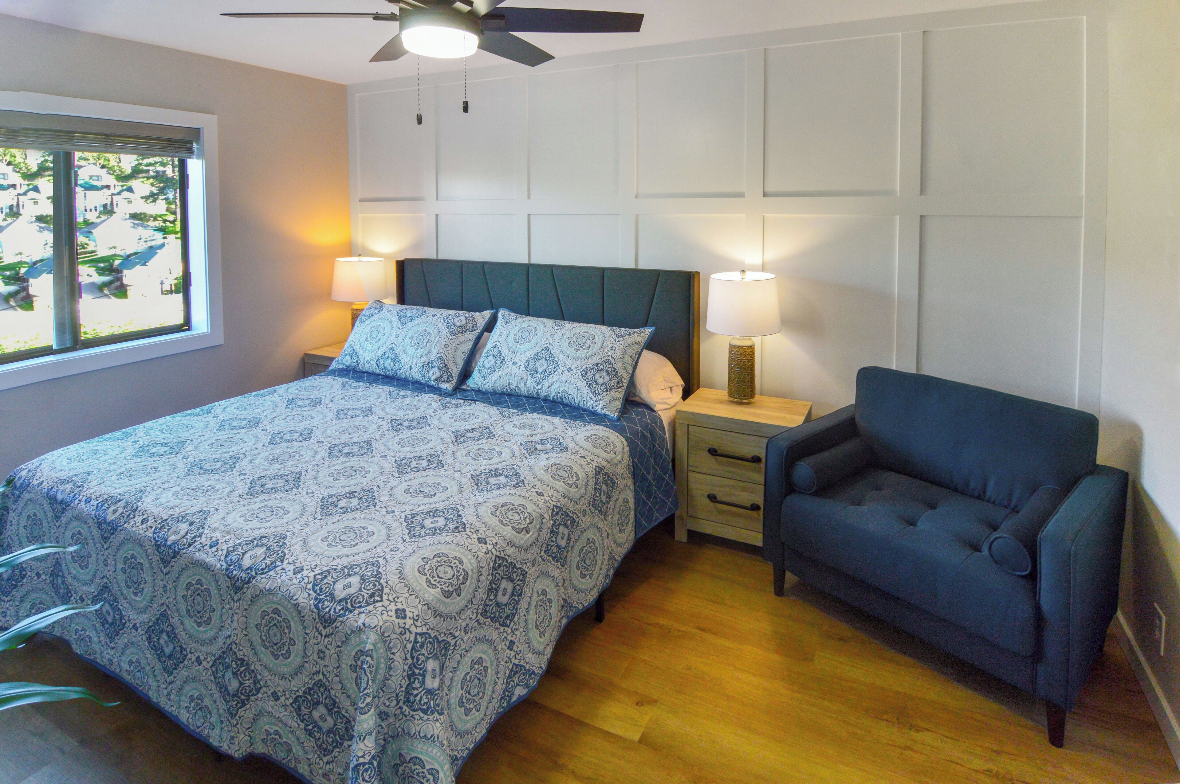 Een slaapkamer met een bed, nachtkastjes, een stoel, een plafondventilator en een raam met uitzicht op huizen.