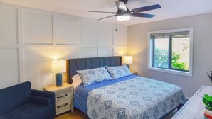 A bedroom with a bed, bedside tables, a ceiling fan, a window with a view, and a small table with a plant.