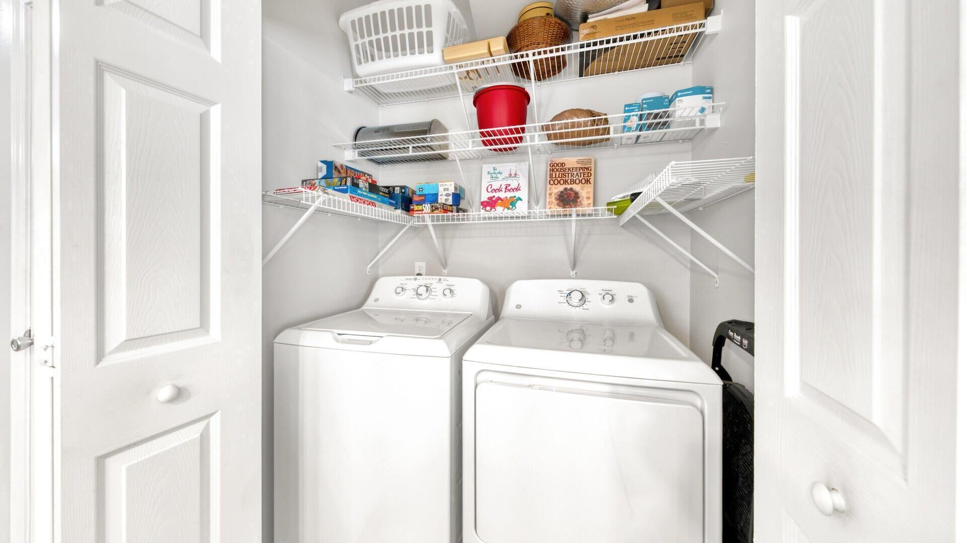 A laundry room with two white front-loading washers and dryers, shelves above holding various items including a red cup, baskets, and boxes.