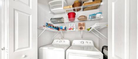 A laundry room with two white front-loading washers and dryers, shelves above holding various items including a red cup, baskets, and boxes.