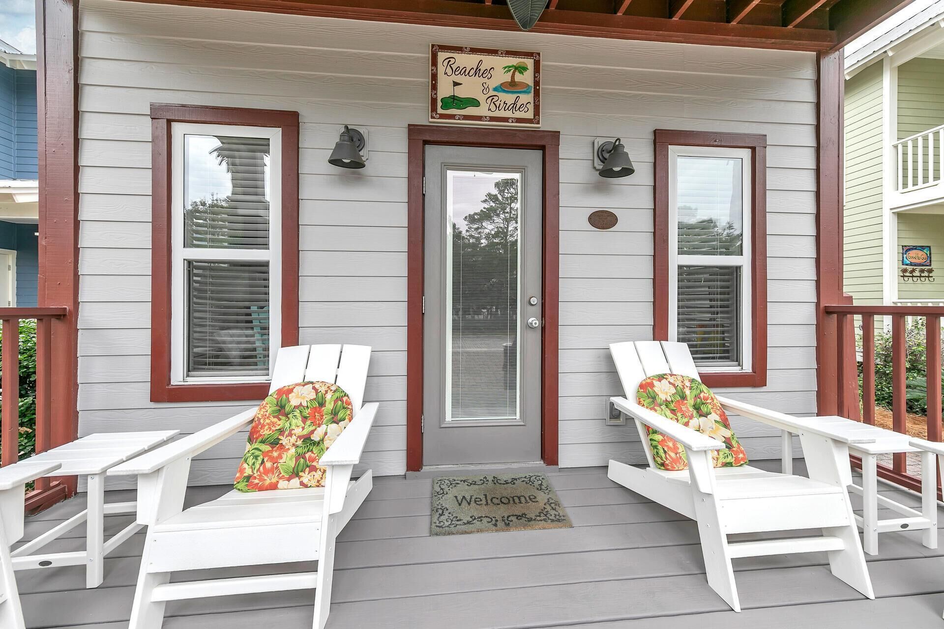 Eine Veranda mit zwei weißen Adirondack-Stühlen, einer Fußmatte und einem Schild mit der Aufschrift 'Beaches & Birds'.