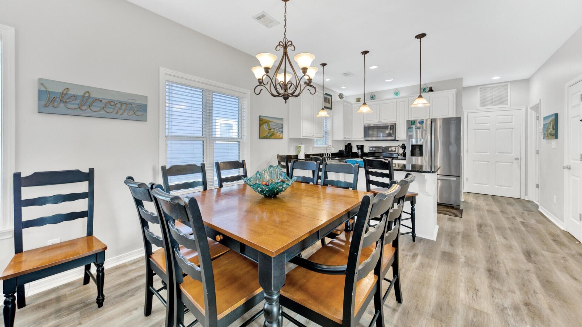 A dining area with a wooden table and chairs, a chandelier, and a kitchen in the background.