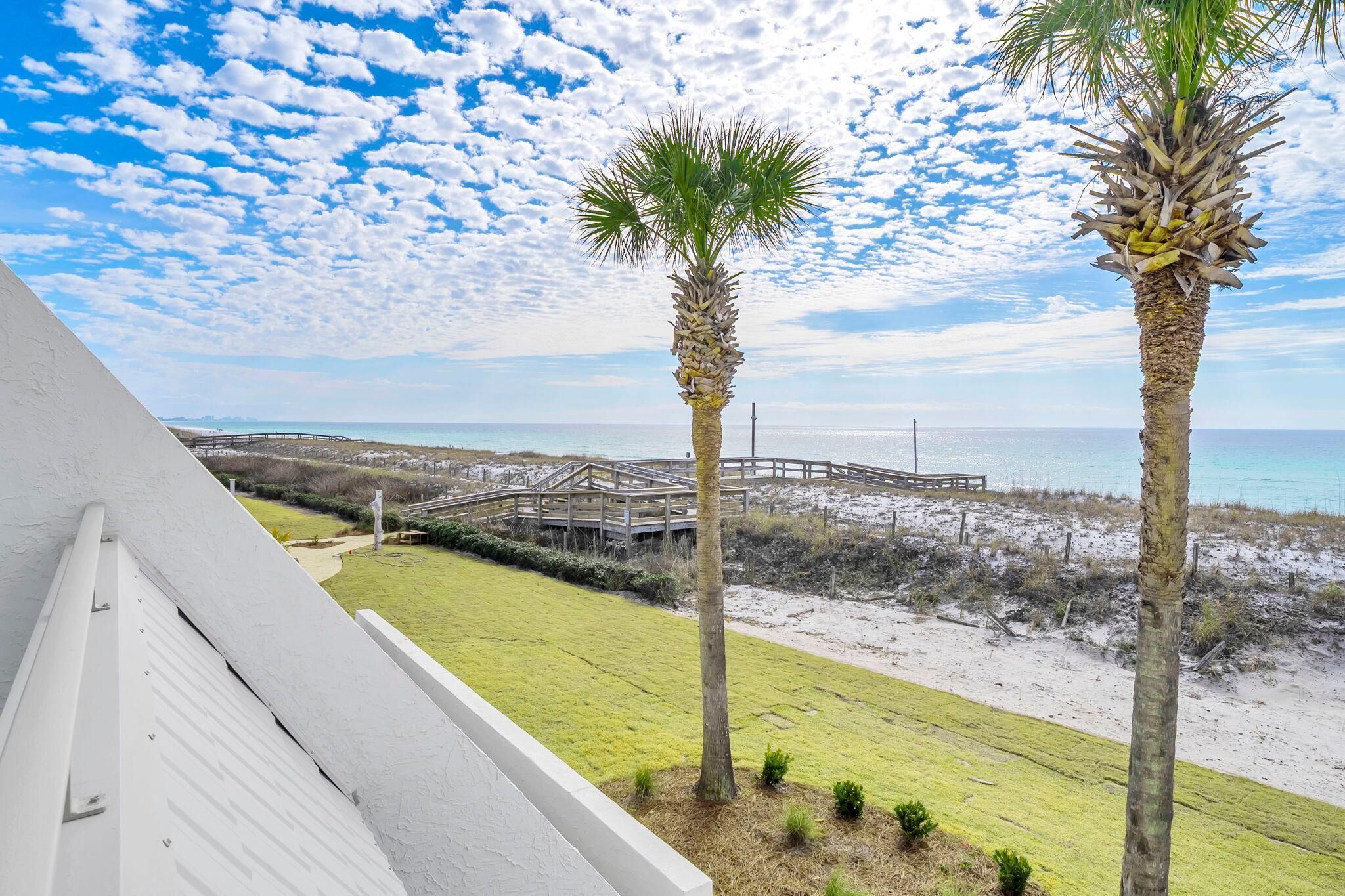 Une vue depuis un balcon donnant sur une plage bordée de palmiers, avec un pont en bois et un ciel dégagé.