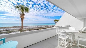 Un balcon avec une table et des chaises blanches, un palmier, et une vue sur la plage et l’océan.