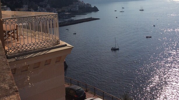 A coastal view with boats on the water, a mountainous backdrop, and a building with a balcony.