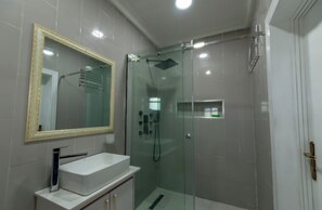 A modern bathroom with a glass-enclosed shower, a white sink with a gold-framed mirror, and tiled walls. - De Grand Hotel and Rooftop (Calabar)
