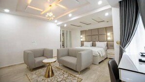 A modern hotel room with a large bed, two armchairs, a small round table, and a decorative ceiling light. - De Grand Hotel and Rooftop (Calabar)