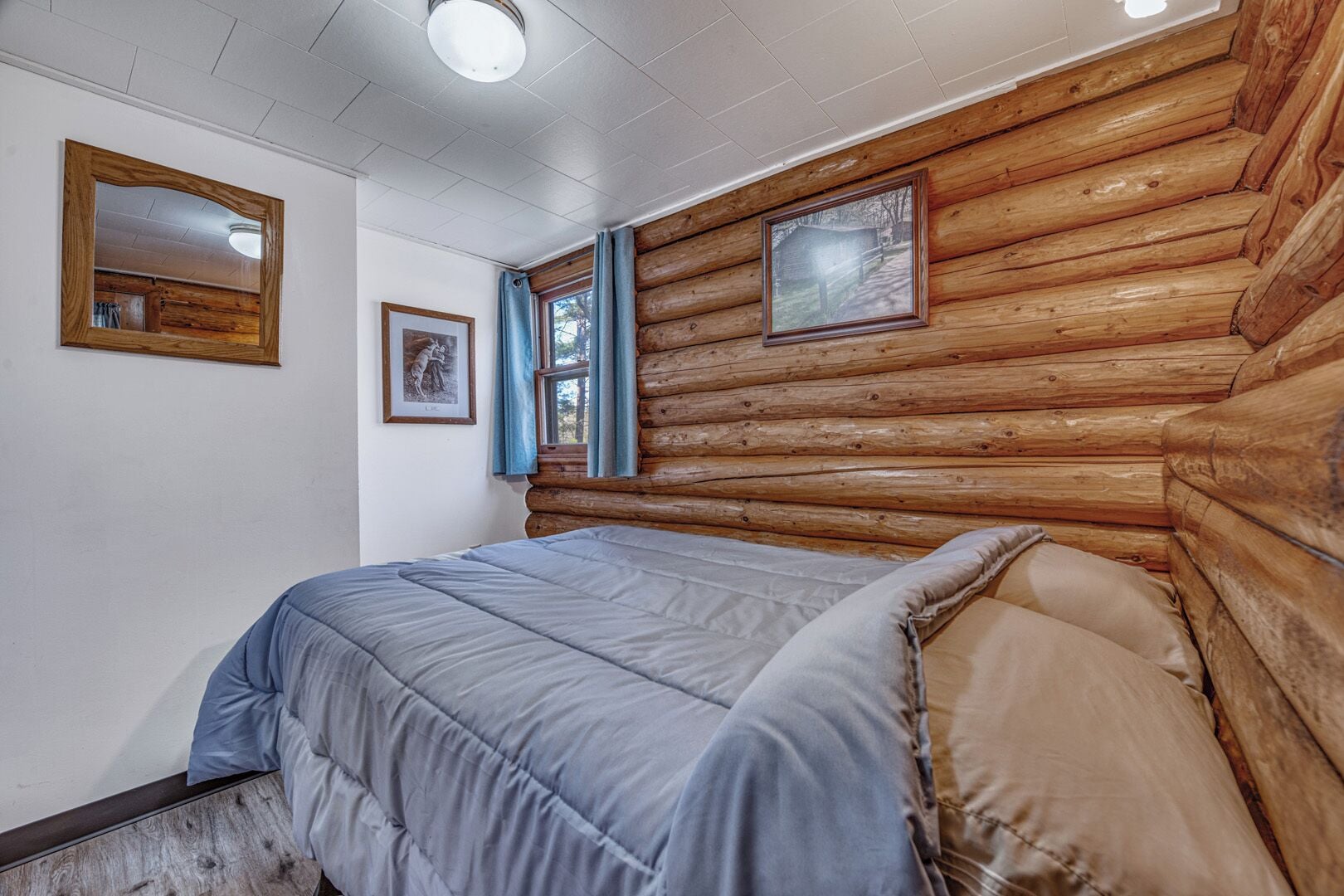 A log cabin bedroom with a bed, wooden walls, a window with curtains, and framed pictures.