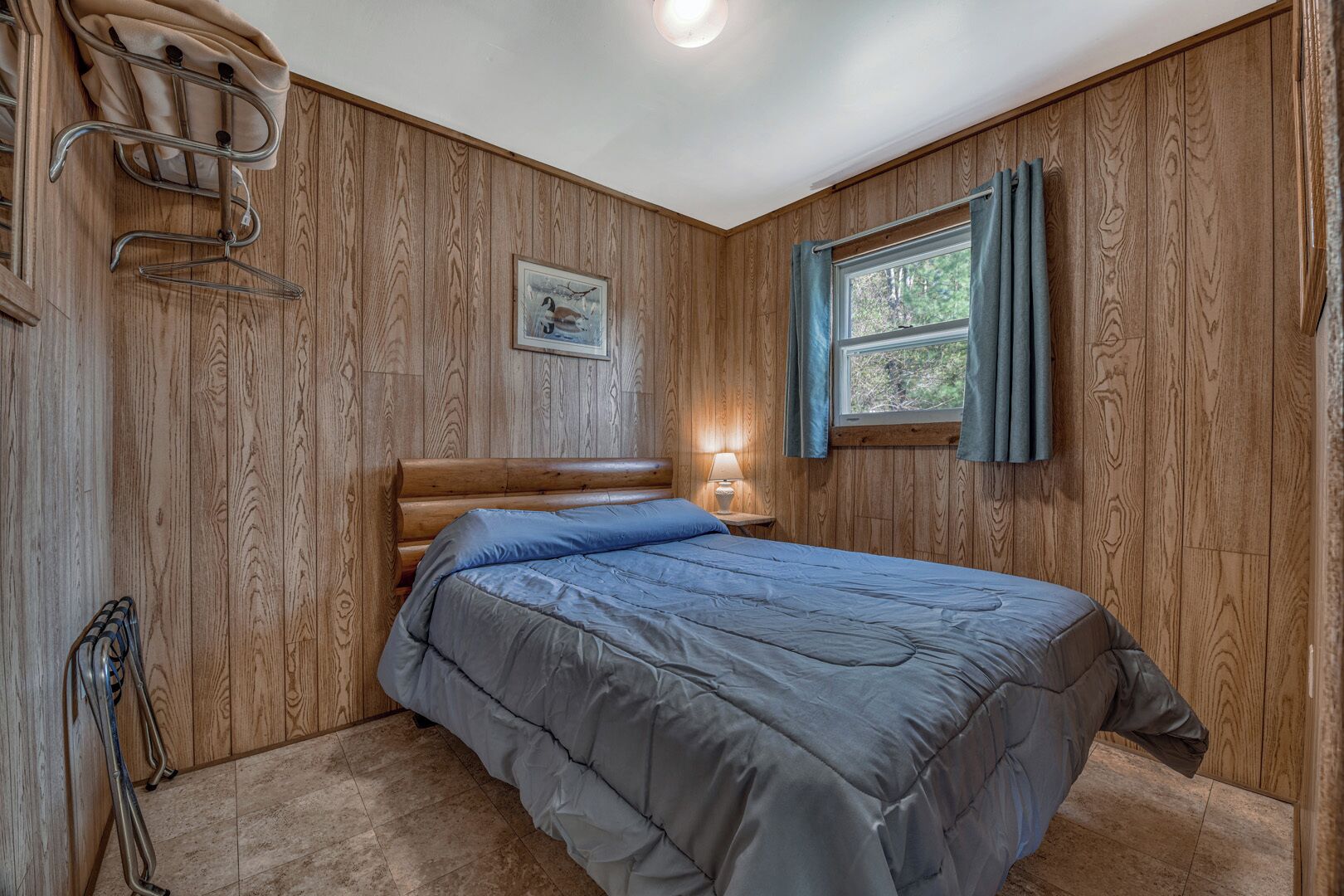 Ein Schlafzimmer mit einem hölzernen Kopfteil, einem Bett mit grauer Bettdecke, einer Nachttischlampe und einem Fenster mit Vorhängen.