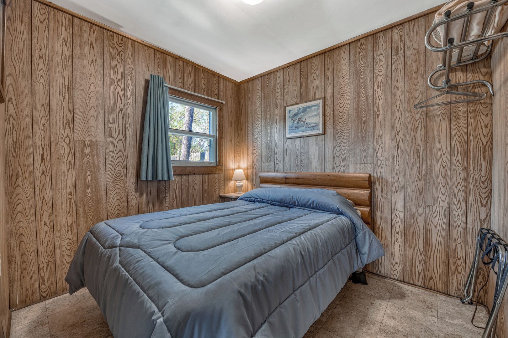 Un dormitorio con pared de paneles de madera, una cama con cubrecama azul, una ventana con cortinas, una lámpara de noche y un radiador.