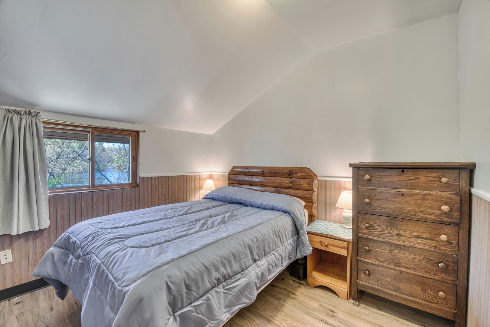 Un dormitorio con cama, cómoda de madera y ventana con vistas a árboles.