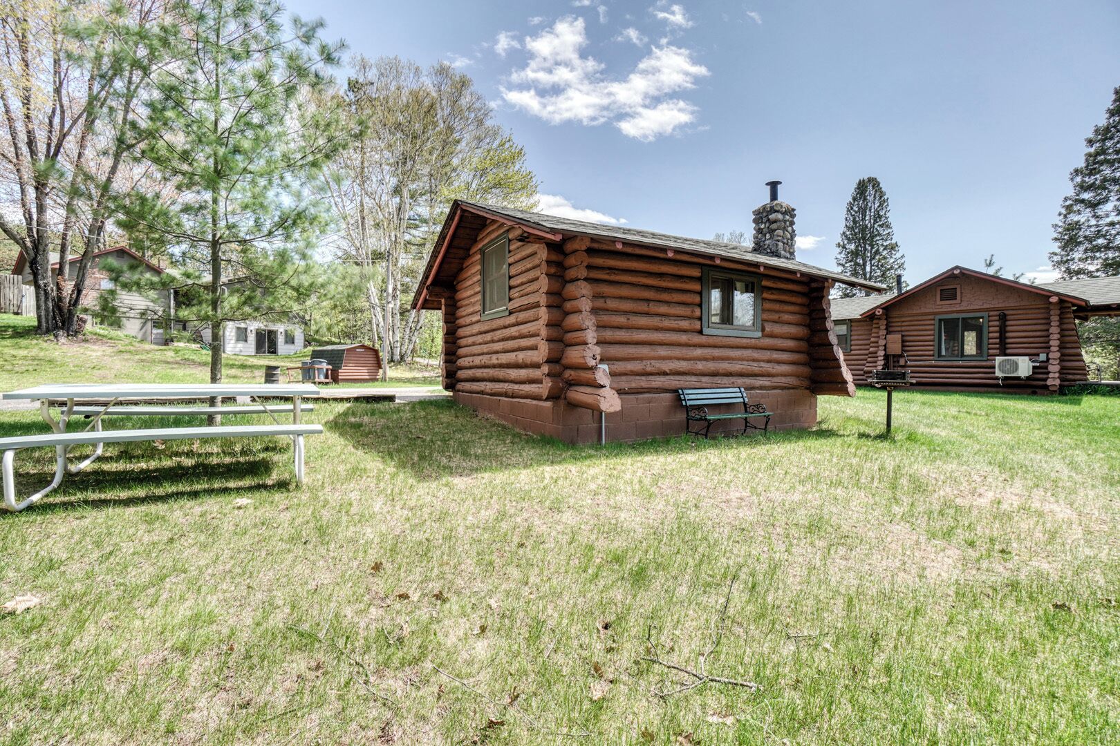 A rustic log cabin with a chimney, a picnic table, and a bench in a grassy area with trees.