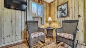 A room with wooden paneled walls, a flat-screen TV mounted on the wall, two wicker chairs with striped cushions, a small table with a lamp, and a framed picture on the wall. - Edgewater Resort (Iron Mountain)