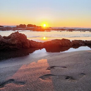A beach scene with a sunset, rocks, and calm water reflecting the sun.