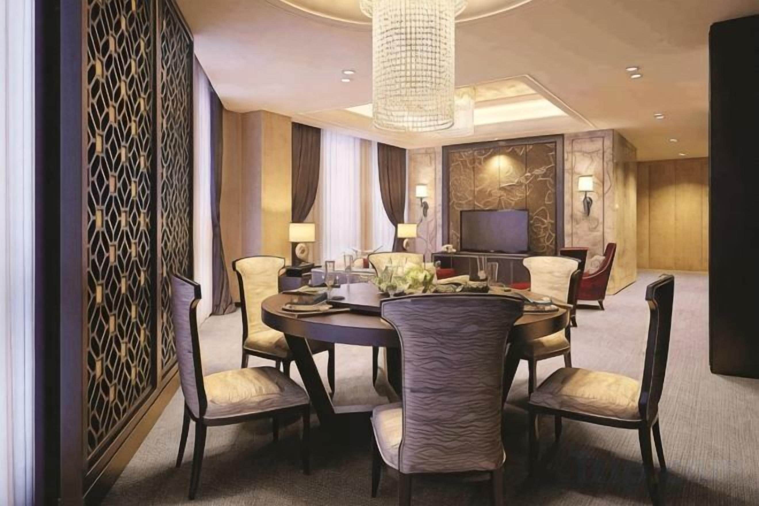 A hotel dining room with a round table, chairs, a chandelier, and a television.