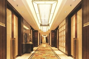 Un long couloir étroit d’hôtel, avec des lambris en bois, une moquette à motifs et un lustre central.